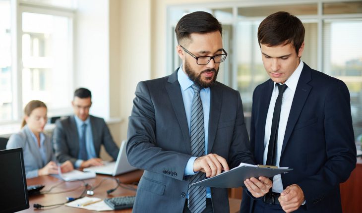 Confident businessman pointing at document while explaining his idea to his partner on background of their colleagues